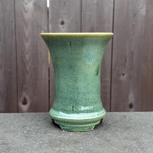 May include: A handcrafted, green ceramic vase with a speckled texture. The vase has a flared top and a rounded base, with a light green rim. The vase is approximately 7 inches tall, perfect for displaying flowers or as a decorative piece.