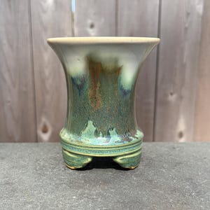 May include: A handcrafted ceramic vase with a flared top and a pedestal base. The vase features a blend of green, blue, and brown glaze, creating a unique, textured appearance. The vase is supported by three small feet.