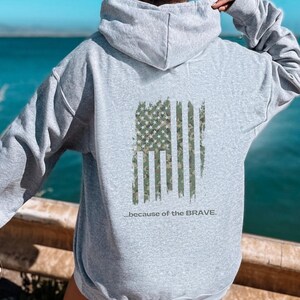 Może przedstawiać: Szara bluza z kapturem z przetartym wzorem amerykańskiej flagi z kamuflażem. Pod flagą nadrukowany jest tekst "...because of the BRAVE."