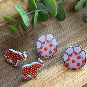 May include: A collection of four decorative pins. Two pins are shaped like elephants with red polka dots on a white background. The other two pins are paw prints with red polka dots on a white background. The pins are on a wooden surface.