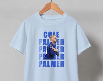 Op de afbeelding: Een lichtblauw t-shirt met korte mouwen en een grafische print van een voetballer in een blauw tenue die een doelpunt viert. De tekst COLE PALMER staat verticaal herhaald in blauwe blokletters achter de speler op de voorkant van het shirt.