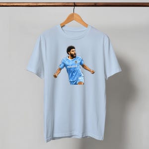 May include: Light blue t-shirt featuring a graphic of a soccer player in a blue jersey with the word "monzo" on the front. The player is celebrating with arms raised. The shirt is hanging on a wooden hanger.
