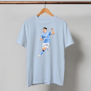 May include: Light blue t-shirt featuring a full-color image of a soccer player in a light blue uniform. The player is running and celebrating. The shirt is hanging on a wooden hanger against a light blue background.