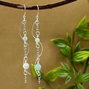 Könnte beinhalten: Ein Paar Ohrhänger mit silberfarbenem Metalldesign. Jeder Ohrring hat eine geschwungene, offene Form mit einer Kette aus kleinen Gliedern und drei hellblauen Perlen. Die Ohrringe hängen an einem geschwungenen Ast vor neutralem Hintergrund.