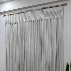 以下が含まれることがあります： 織り模様とフリンジが付いた白いマクラメの壁掛け。ナチュラルなコットンコードで作られており、木製の棒に取り付けられています。