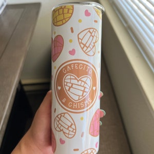 May include: White stainless steel tumbler with a white background and a pattern of pink and yellow heart-shaped pastries. The tumbler has a black circle with the text "Cafecito & Chisme" in white.