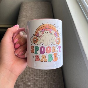 Puede incluir: Taza de cer&aacute;mica blanca con asa e interior rosa. La taza presenta un dise&ntilde;o colorido con un fantasma con sombrero de bruja, un arco&iacute;ris y las palabras "SPOOKY BABE" en una fuente retro. El dise&ntilde;o incluye flores y murci&eacute;lagos.
