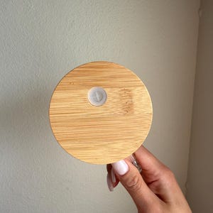 K&ouml;nnte beinhalten: Ein runder, hellbrauner Bambusdeckel mit einem wei&szlig;en Kunststoffverschluss. Der Deckel wird von einer Hand mit manik&uuml;rten N&auml;geln gehalten. Der Bambus hat ein nat&uuml;rliches Holzmaserungsmuster. Der Deckel ist f&uuml;r ein Glas oder eine Flasche konzipiert.