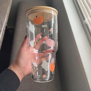 May include: A clear glass tumbler with a bamboo lid, decorated with orange pumpkins, white ghosts, and crescent moons. The tumbler is designed with a Halloween theme, perfect for seasonal beverages. The glass is held by a person.