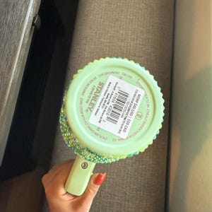 May include: A light green Stanley Flowstate tumbler with a handle, embellished with iridescent rhinestones around the rim. The tumbler has a matching green handle and a lid. The bottom of the tumbler has product information printed on it.