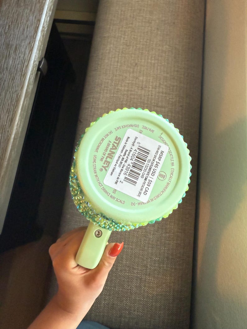 May include: A light green Stanley Flowstate tumbler with a handle, embellished with iridescent rhinestones around the rim. The tumbler has a matching green handle and a lid. The bottom of the tumbler has product information printed on it.