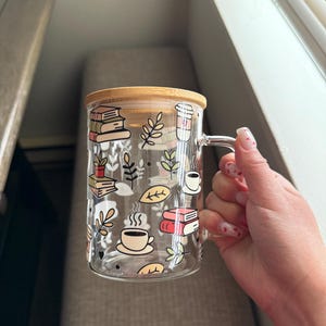 Op de afbeelding: Een heldere glazen mok met een bamboe deksel heeft een speels ontwerp met boeken, koffiekopjes en bladeren. De mok is perfect om van een warme drank te genieten.