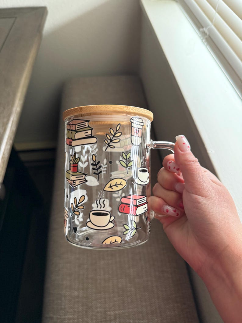Op de afbeelding: Een heldere glazen mok met een bamboe deksel heeft een speels ontwerp met boeken, koffiekopjes en bladeren. De mok is perfect om van een warme drank te genieten.