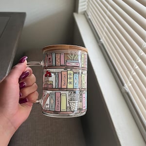 May include: A glass mug with a wooden lid features a colorful illustration of a bookshelf filled with books. The books have titles like "Rainy Day", "Romantic", "Spice", and "Book Lover".