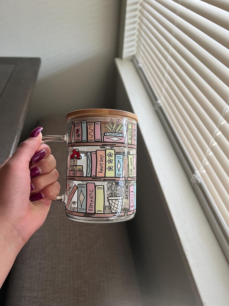 May include: A glass mug with a wooden lid features a colorful illustration of a bookshelf filled with books. The books have titles like "Rainy Day", "Romantic", "Spice", and "Book Lover".