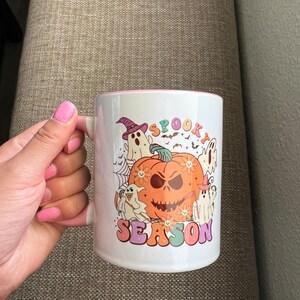 Puede incluir: Taza de cer&aacute;mica blanca con interior rosa, con un dise&ntilde;o tem&aacute;tico de Halloween. El dise&ntilde;o incluye una calabaza, fantasmas, murci&eacute;lagos y las palabras "Spooky Season" en una fuente retro. La taza tiene un asa blanca.