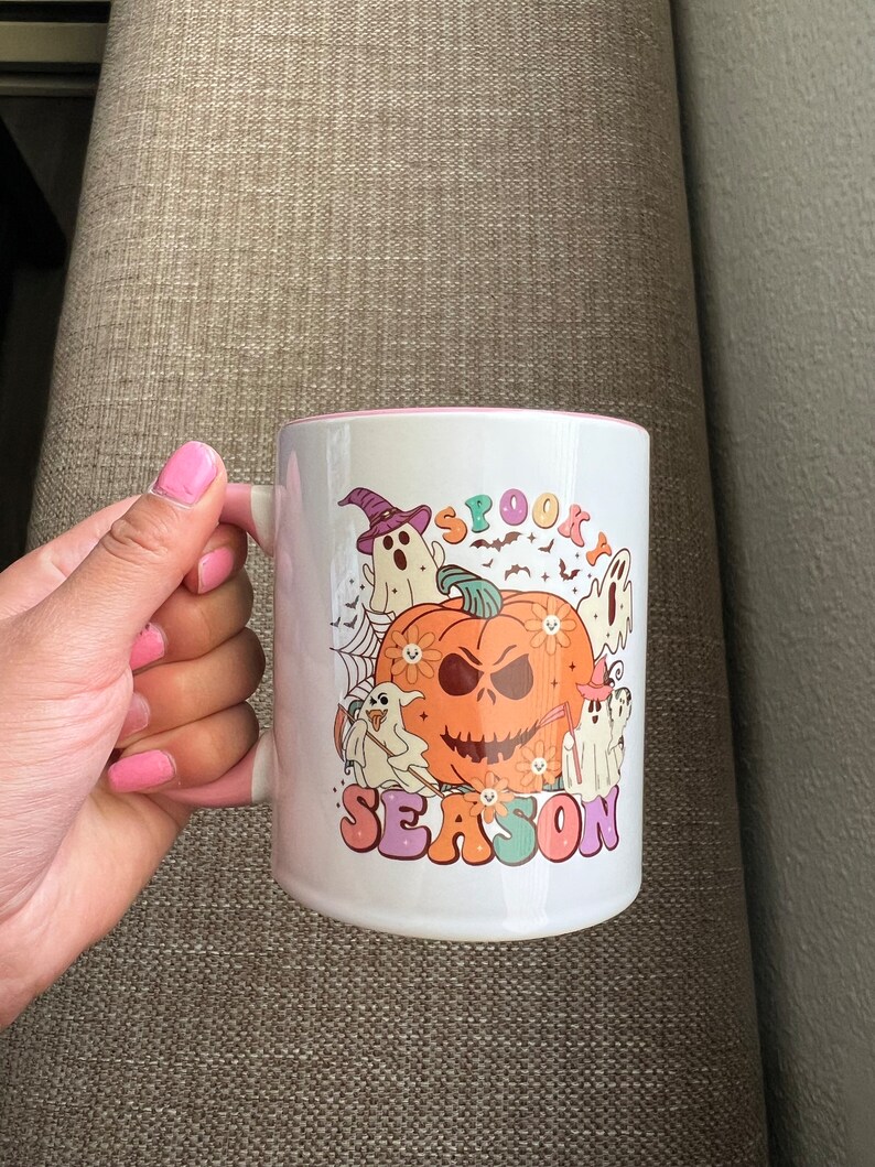Puede incluir: Taza de cer&aacute;mica blanca con interior rosa, con un dise&ntilde;o tem&aacute;tico de Halloween. El dise&ntilde;o incluye una calabaza, fantasmas, murci&eacute;lagos y las palabras "Spooky Season" en una fuente retro. La taza tiene un asa blanca.