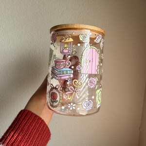 K&ouml;nnte beinhalten: Ein klares Glas mit Bambusdeckel, verziert mit pastellfarbenen Illustrationen. Das Design umfasst eine Laterne, eine Teetasse, B&uuml;cher, eine T&uuml;r, Rosen und Schmetterlinge. Die Dose wird von einer Person gehalten, die einen roten Pullover tr&auml;gt.
