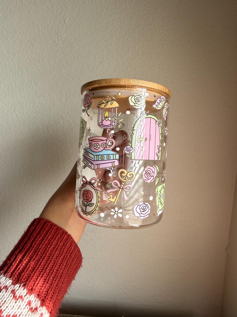 K&ouml;nnte beinhalten: Ein klares Glas mit Bambusdeckel, verziert mit pastellfarbenen Illustrationen. Das Design umfasst eine Laterne, eine Teetasse, B&uuml;cher, eine T&uuml;r, Rosen und Schmetterlinge. Die Dose wird von einer Person gehalten, die einen roten Pullover tr&auml;gt.
