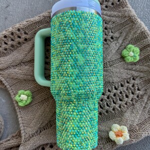 May include: A green and teal bedazzled tumbler with a handle and a straw. The tumbler is covered in sparkling rhinestones. The lid and handle are a matching light green color. The tumbler is resting on a brown knitted surface with three small flower accents.