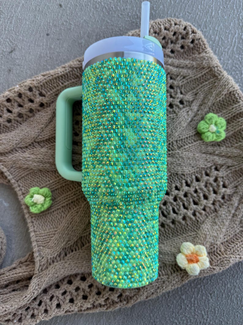 May include: A green and teal bedazzled tumbler with a handle and a straw. The tumbler is covered in sparkling rhinestones. The lid and handle are a matching light green color. The tumbler is resting on a brown knitted surface with three small flower accents.