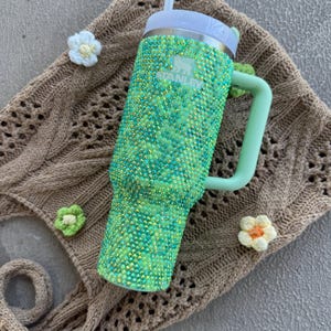 May include: A green and blue bedazzled Stanley tumbler with a white lid and straw. The tumbler has a light green handle and the Stanley logo. The tumbler is on a brown crocheted bag with white and green crocheted flowers.