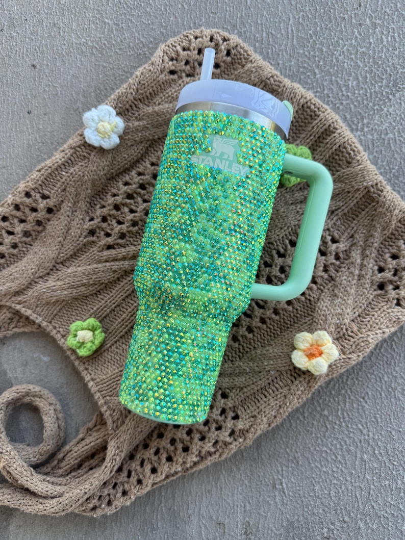 May include: A green and blue bedazzled Stanley tumbler with a white lid and straw. The tumbler has a light green handle and the Stanley logo. The tumbler is on a brown crocheted bag with white and green crocheted flowers.