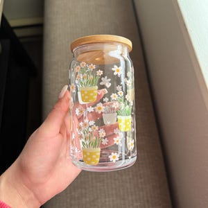 May include: A clear glass jar with a wooden lid, decorated with a floral pattern of white daisies and yellow potted plants.