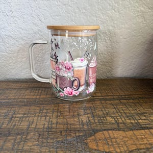 May include: A clear glass coffee mug with a wooden lid. The mug features a floral and coffee-themed design with the words "Coffee first!" printed on it. The design includes images of coffee drinks, flowers, and butterflies.