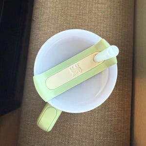 May include: A white sippy cup with a light green lid and handle. The lid has a light green band with a white rectangle and a small design. A white straw extends from the lid.