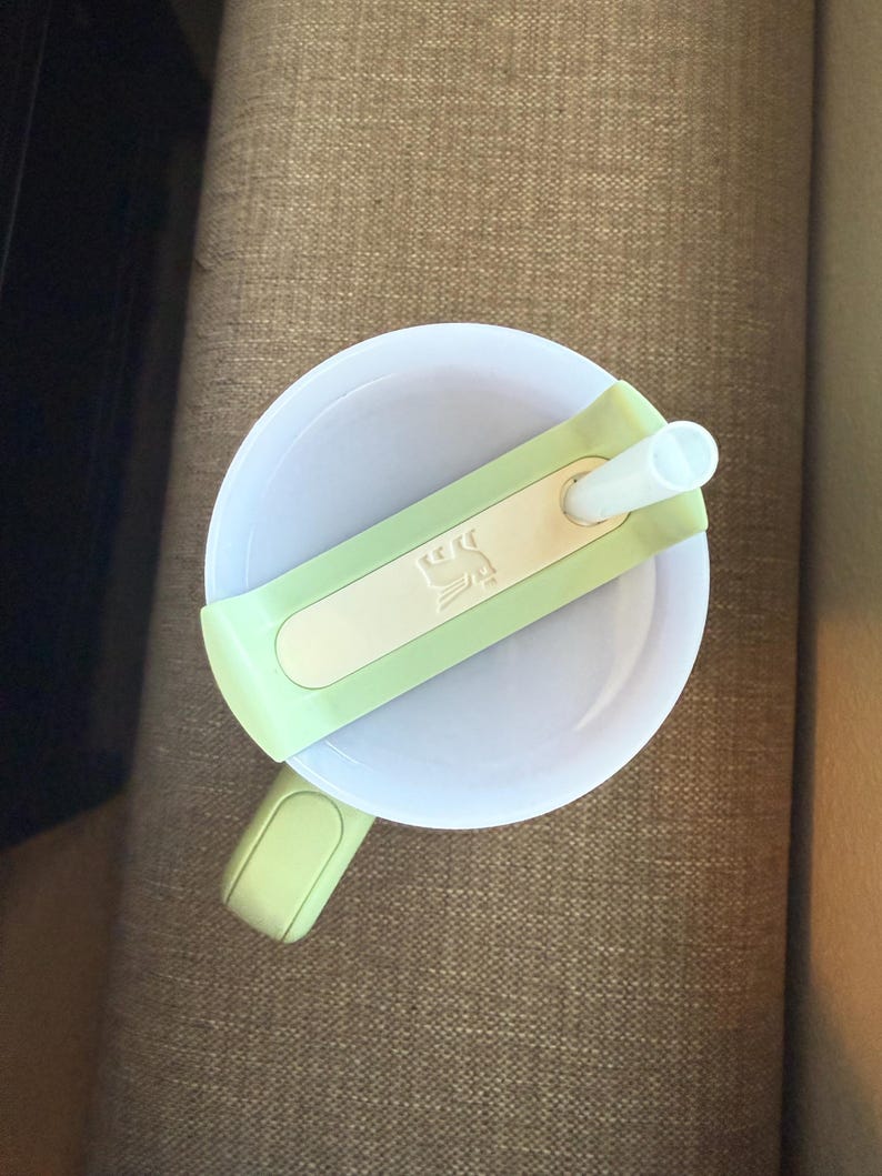 May include: A white sippy cup with a light green lid and handle. The lid has a light green band with a white rectangle and a small design. A white straw extends from the lid.