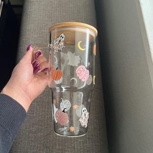 May include: A clear glass tumbler with a wooden lid, featuring Halloween-themed designs. The tumbler has a handle and is decorated with orange and pink pumpkins, ghosts, and crescent moons. The tumbler is held by a person.