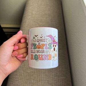 Puede incluir: Taza de cer&aacute;mica blanca con interior rosa, con un dise&ntilde;o colorido con un fantasma con sombrero de bruja, una calabaza y el texto "I ghost people all year round". La taza tiene un asa y la sostiene una persona.