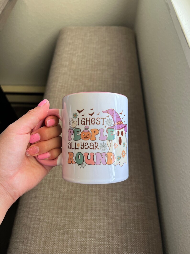 Puede incluir: Taza de cer&aacute;mica blanca con interior rosa, con un dise&ntilde;o colorido con un fantasma con sombrero de bruja, una calabaza y el texto "I ghost people all year round". La taza tiene un asa y la sostiene una persona.
