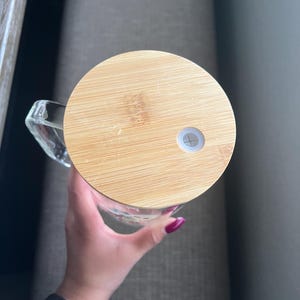 May include: A clear glass mug with a handle, topped with a round, light brown bamboo lid. The lid has a small white plug. The mug is held by a person.