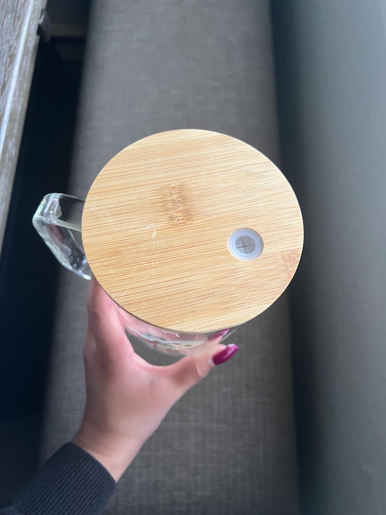 May include: A clear glass mug with a handle, topped with a round, light brown bamboo lid. The lid has a small white plug. The mug is held by a person.