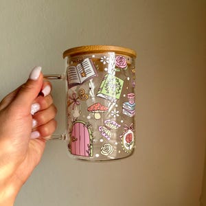 K&ouml;nnte beinhalten: Transparente Glas-Tasse mit Bambusdeckel, verziert mit skurrilen Illustrationen. Das Design umfasst eine rosa T&uuml;r, B&uuml;cher, Pilze und Rosen. Die Tasse hat einen transparenten Griff.