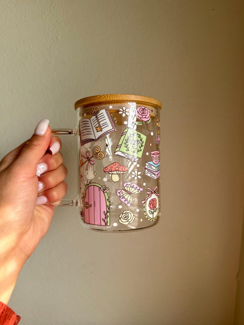 K&ouml;nnte beinhalten: Transparente Glas-Tasse mit Bambusdeckel, verziert mit skurrilen Illustrationen. Das Design umfasst eine rosa T&uuml;r, B&uuml;cher, Pilze und Rosen. Die Tasse hat einen transparenten Griff.