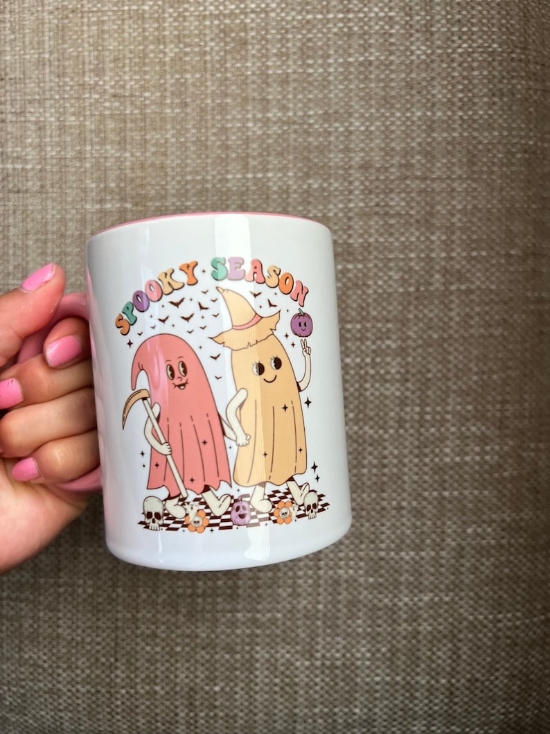 Puede incluir: Taza de cer&aacute;mica blanca con interior y asa rosas. La taza presenta una colorida ilustraci&oacute;n de dos fantasmas de dibujos animados con el texto "Spooky Season". Un fantasma sostiene una guada&ntilde;a y el otro un orbe morado.