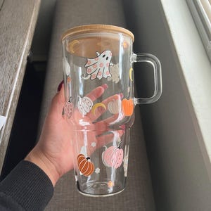 May include: A clear glass mug with a wooden lid and handle, decorated with Halloween-themed illustrations. The design includes pumpkins, ghosts, and crescent moons in orange, pink, and white. The mug is designed to hold a beverage.