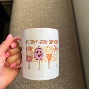 Puede incluir: Taza de cer&aacute;mica blanca con asa y borde rosa. La taza presenta el texto "SWEET AND SPOOKY" en letras naranjas, junto con ilustraciones de golosinas tem&aacute;ticas de Halloween, como calabazas y fantasmas en palitos. La taza es sostenida por una persona con esmalte de u&ntilde;as rosa.