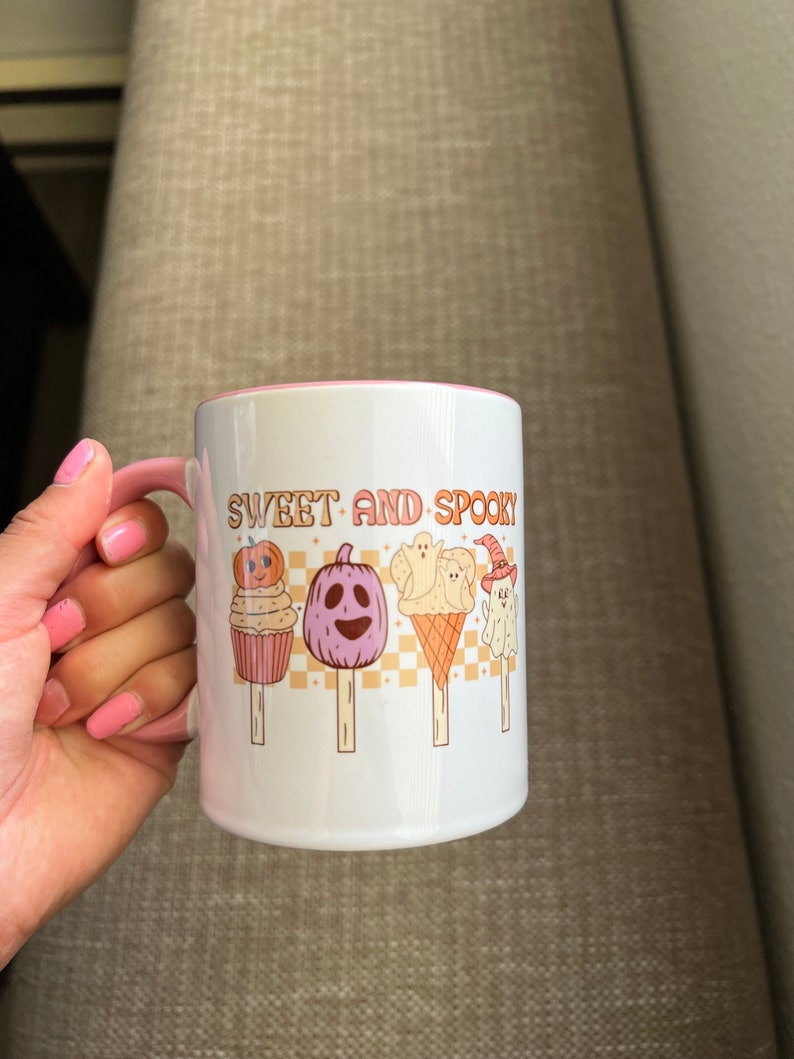 Puede incluir: Taza de cer&aacute;mica blanca con asa y borde rosa. La taza presenta el texto "SWEET AND SPOOKY" en letras naranjas, junto con ilustraciones de golosinas tem&aacute;ticas de Halloween, como calabazas y fantasmas en palitos. La taza es sostenida por una persona con esmalte de u&ntilde;as rosa.