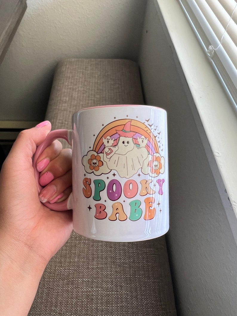 Puede incluir: Taza de cer&aacute;mica blanca con asa e interior rosa. La taza presenta un dise&ntilde;o colorido con un fantasma con sombrero de bruja, un arco&iacute;ris y las palabras "SPOOKY BABE" en una fuente retro. El dise&ntilde;o incluye flores y murci&eacute;lagos.