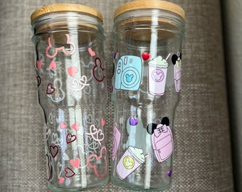 May include: Two glass jars with bamboo lids, decorated with pink and white Mickey Mouse themed designs. The jar on the left has a pink and white Mickey Mouse design with hearts and swirls. The jar on the right has a pink and white Mickey Mouse design with a camera, a cup of coffee, and a backpack. The jars are labeled #1 and #2.