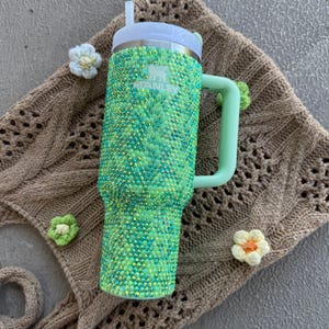 May include: A green and teal bedazzled Stanley tumbler with a white lid and a light green handle. The tumbler has a straw and the Stanley logo. The tumbler is decorated with small, sparkling rhinestones. The tumbler is sitting on a brown knitted surface.
