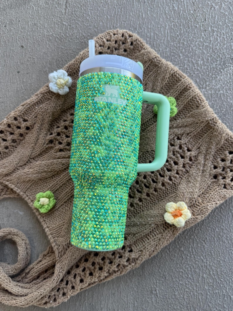 May include: A green and teal bedazzled Stanley tumbler with a white lid and a light green handle. The tumbler has a straw and the Stanley logo. The tumbler is decorated with small, sparkling rhinestones. The tumbler is sitting on a brown knitted surface.