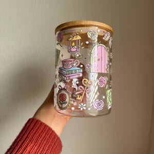 K&ouml;nnte beinhalten: Ein klares Glas mit einem Bambusdeckel, verziert mit skurrilen Illustrationen. Das Glas zeigt Bilder von Rosen, einer rosa T&uuml;r, einem Schl&uuml;ssel, einer Teetasse und einer Laterne. Das Glas hat eine zylindrische Form.