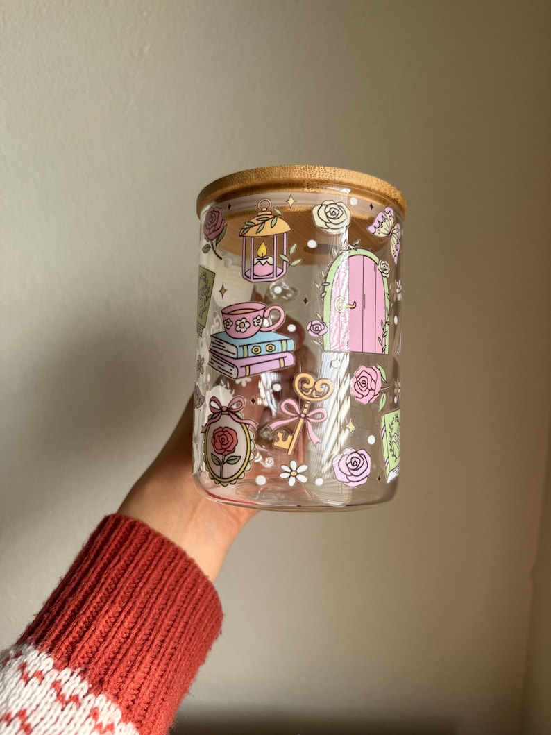 K&ouml;nnte beinhalten: Ein klares Glas mit einem Bambusdeckel, verziert mit skurrilen Illustrationen. Das Glas zeigt Bilder von Rosen, einer rosa T&uuml;r, einem Schl&uuml;ssel, einer Teetasse und einer Laterne. Das Glas hat eine zylindrische Form.