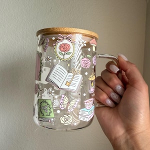 K&ouml;nnte beinhalten: Ein klarer Glasbecher mit Bambusdeckel, verziert mit skurrilen Illustrationen von B&uuml;chern, Rosen, Schmetterlingen und Pilzen. Der Becher hat einen transparenten Griff.