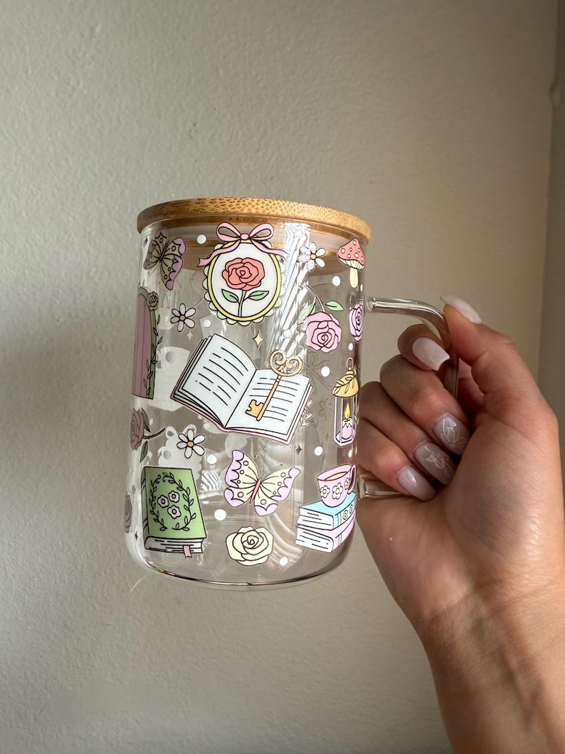 K&ouml;nnte beinhalten: Ein klarer Glasbecher mit Bambusdeckel, verziert mit skurrilen Illustrationen von B&uuml;chern, Rosen, Schmetterlingen und Pilzen. Der Becher hat einen transparenten Griff.